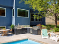 Modern backyard patio with Emory dark outdoor kitchen, pool, dining area, and lush landscaping beside blue-gray house.