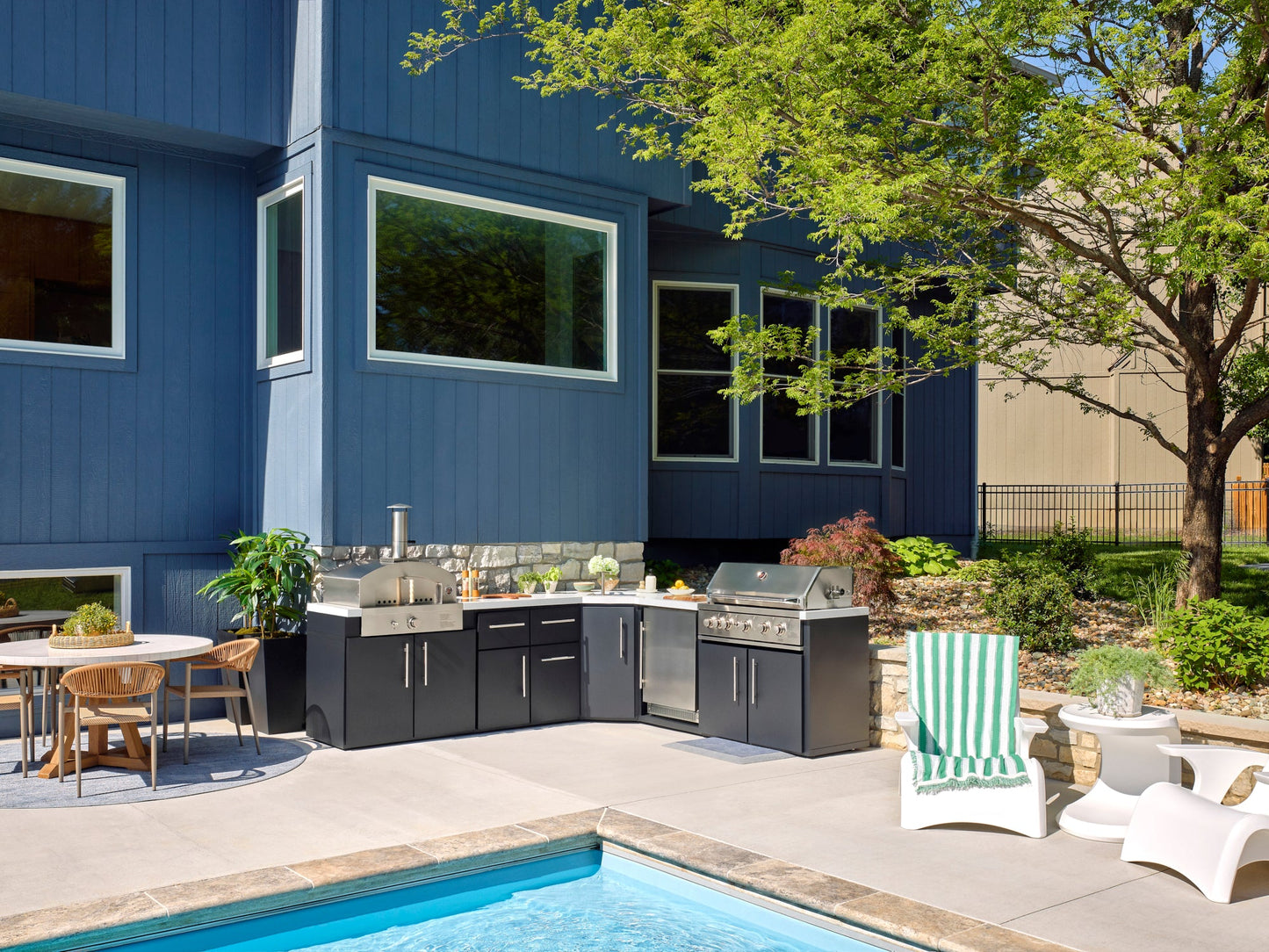 Modern backyard patio with Emory dark outdoor kitchen, pool, dining area, and lush landscaping beside blue-gray house.