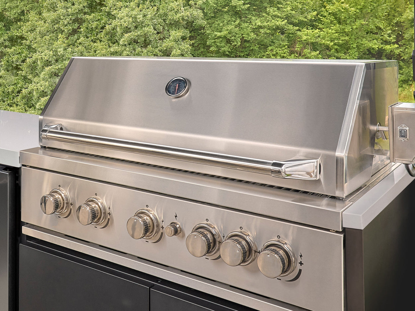 Stainless steel built-in grill with closed lid and temperature gauge, with six control knobs on outdoor patio.
