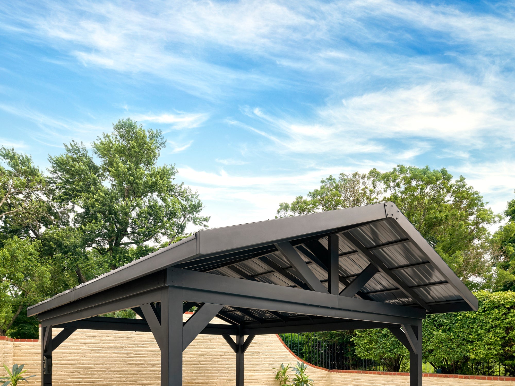 Steel freestanding gazebo with dark metal hardtop roof and exposed beam construction in backyard with trees and fence.