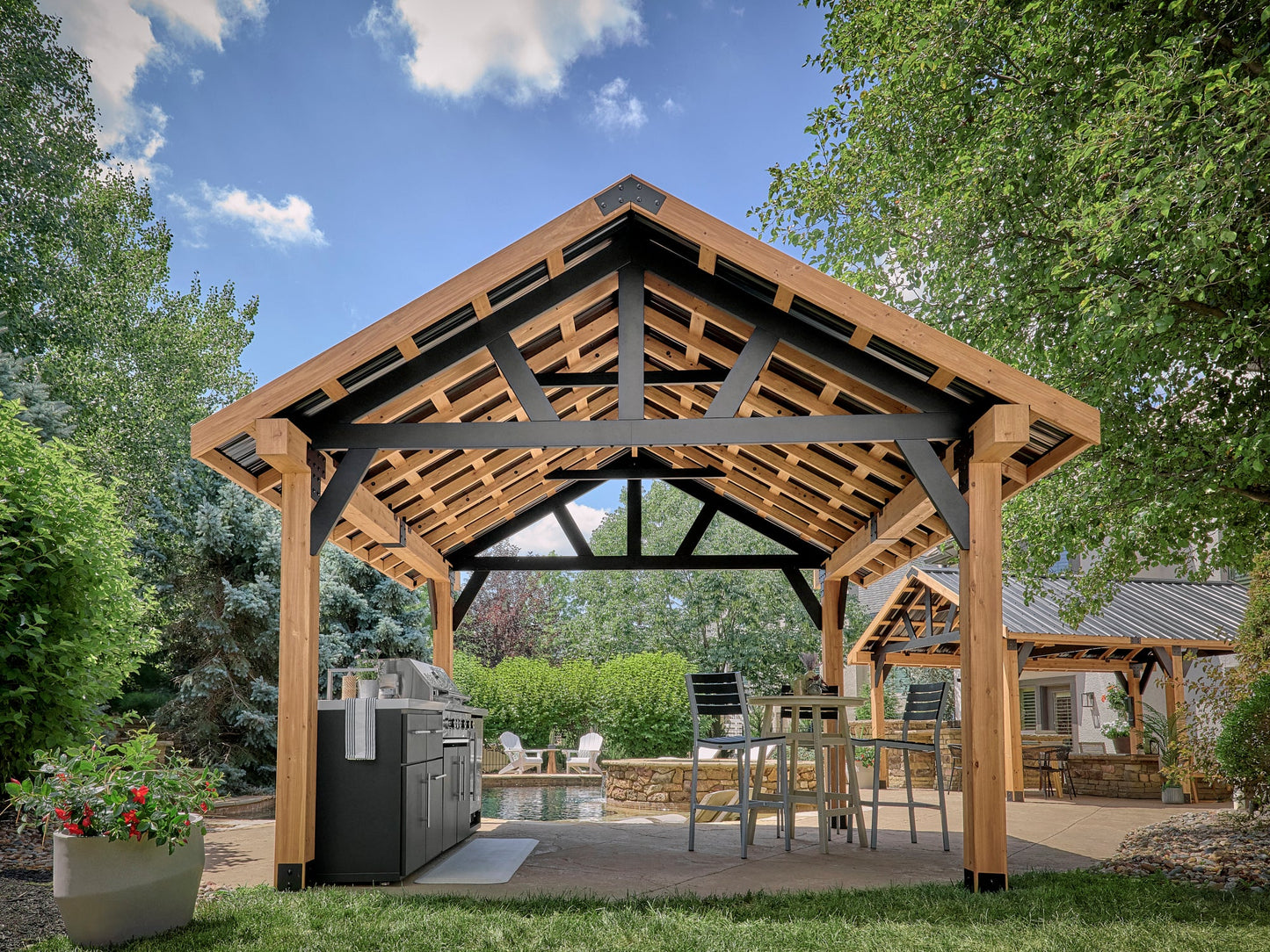 Lodge freestanding gazebo with exposed steel truss roof framing, wooden support posts, outdoor kitchen, and pond.
