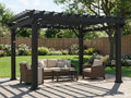 Black metal pergola with open trellis roof and string lights over wicker patio furniture in fenced backyard with gardens.