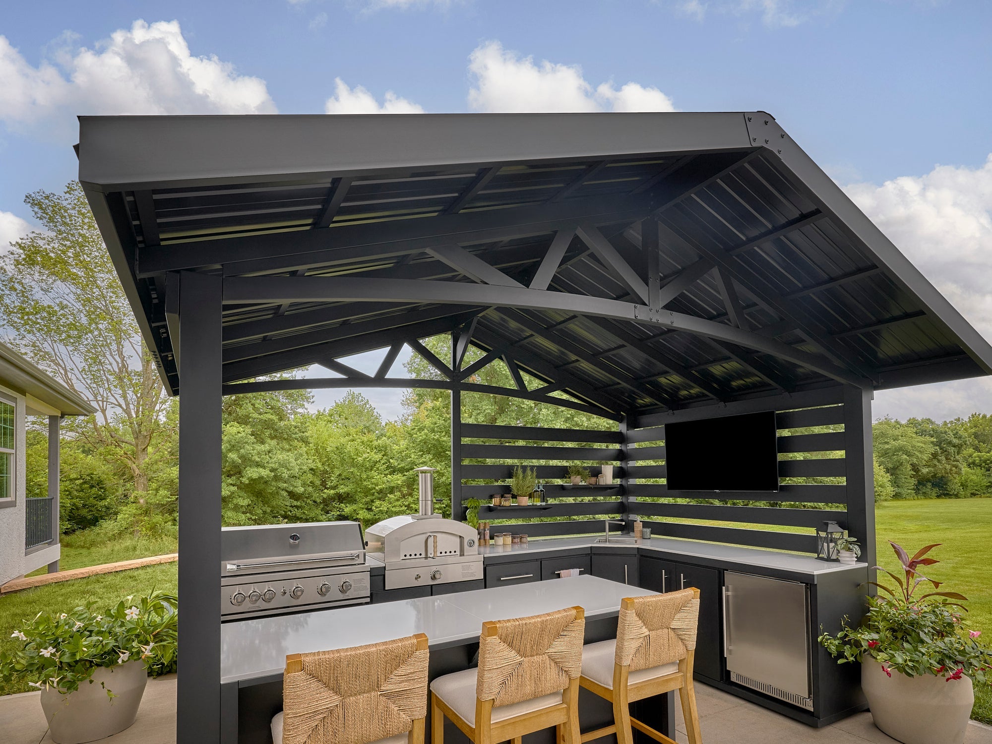 Attached pergola with trellis roof sheltering Langston outdoor kitchen with grill, bar seating, and mounted TV.