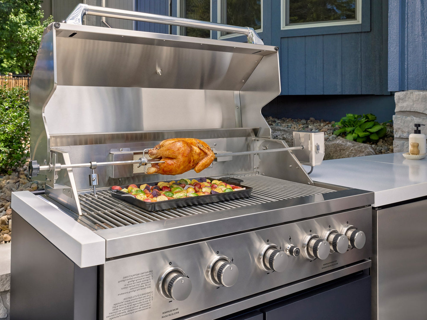 Stainless steel bbq grill with rotisserie chicken and vegetable tray on engineered Emory stone outdoor kitchen countertop.