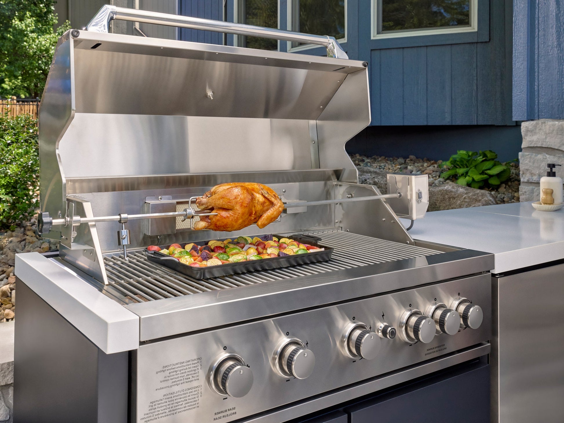Stainless steel bbq grill with rotisserie chicken and vegetable tray on engineered Emory stone outdoor kitchen countertop.