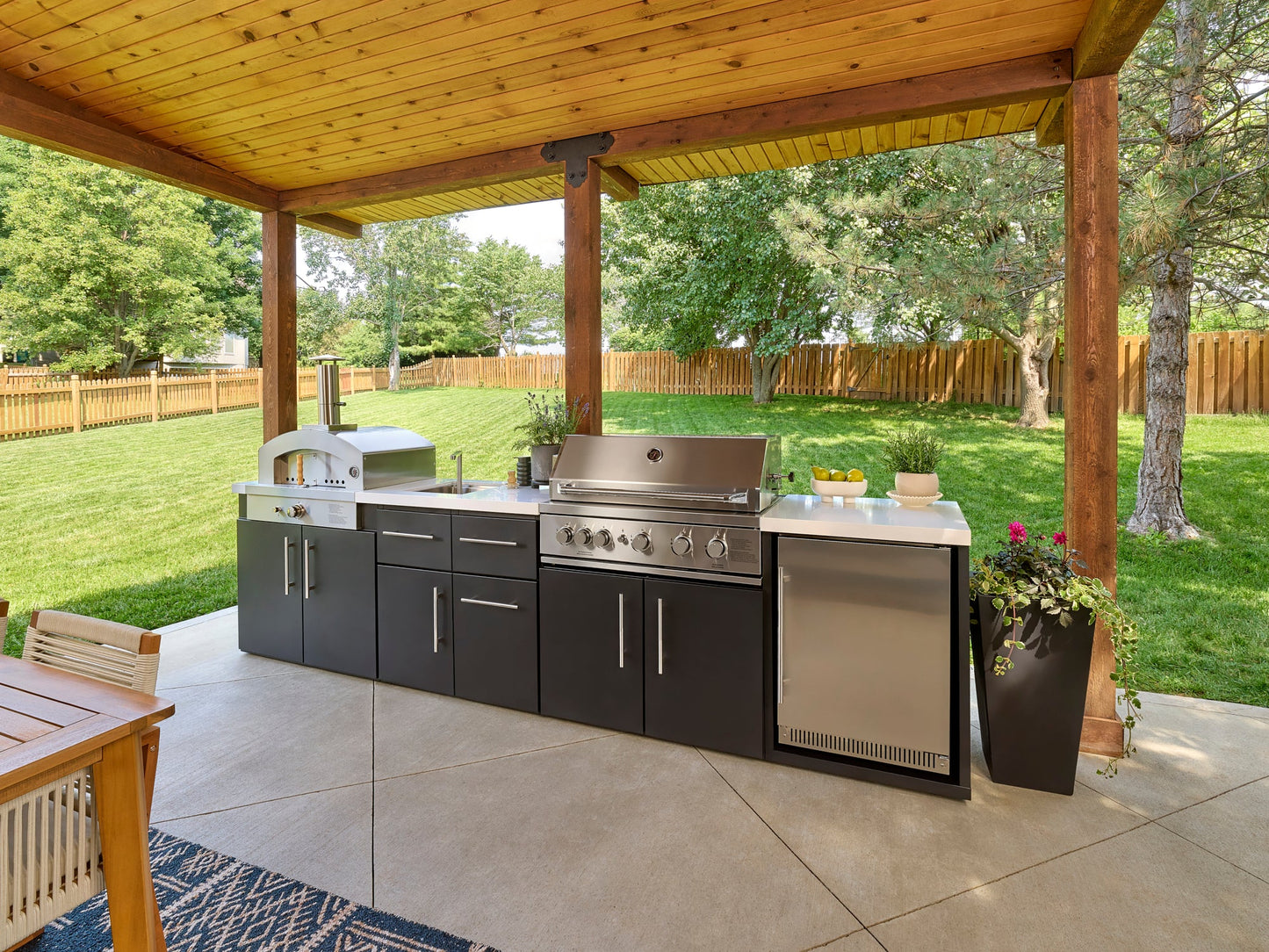 Outdoor cooking station with pizza oven, grill, sink, and refrigerator under covered patio overlooking fenced backyard.